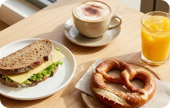 Tisch mit belegtem Vollkornbrot-Sandwich, Laugenbrezel, Cappuccino und Glas Orangensaft