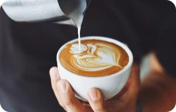Hand hält weiße Tasse mit Latte Art, Milch wird aus kleinem Kännchen eingegossen