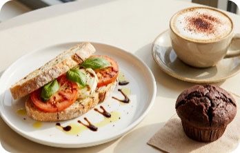 Sandwich mit Tomatenscheiben und Basilikum auf weißem Teller neben braunem Muffin und Cappuccino in Tasse mit Untertasse