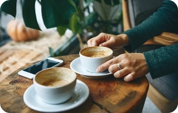 Zwei Tassen Kaffee auf einem Holztisch, eine Hand hält eine Tasse, daneben liegt ein Smartphone