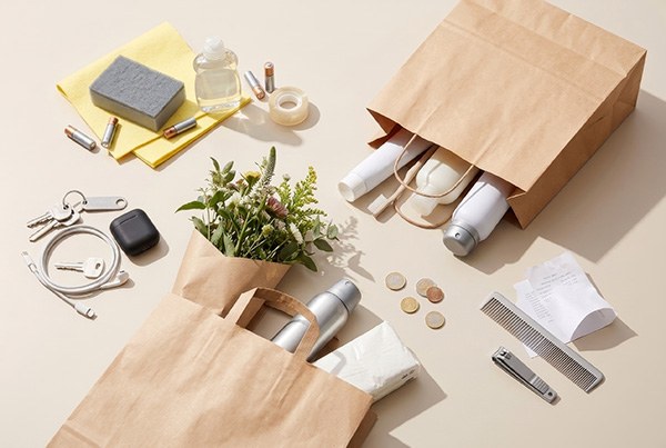 Zwei Papiertüten mit Alltagsgegenständen wie Pflanze, Trinkflasche, Hausschuhe, Nagelknipser, Kamm, Schlüssel, Münzen und Kabel auf hellem Untergrund